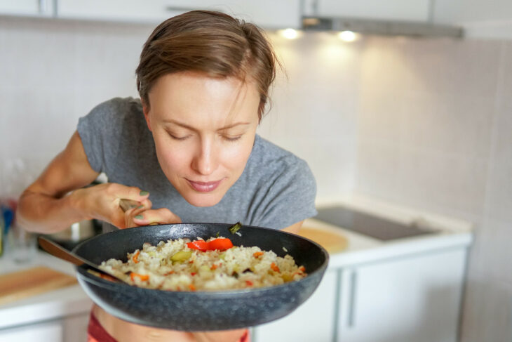 una chica huele la comida que tiene en el sartén que sostiene en la mano