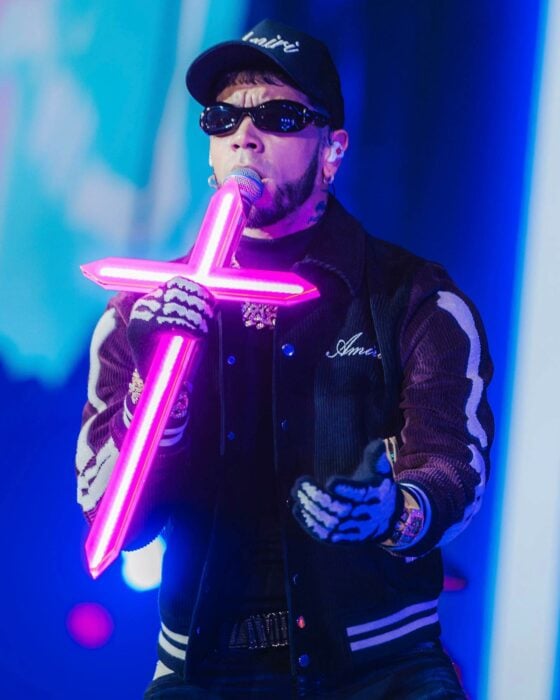 Fotografía de Anuel AA con un micrófono con una cruz iluminada en color rosa 