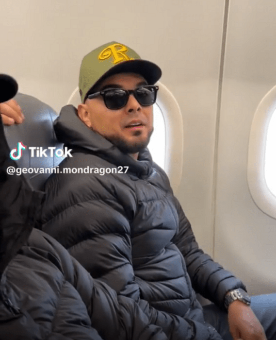 el vocalista de la Banda El Recodo sentado en el asiento de un avión junto a la ventanilla lleva una chamarra oscura gorra y lentes de sol