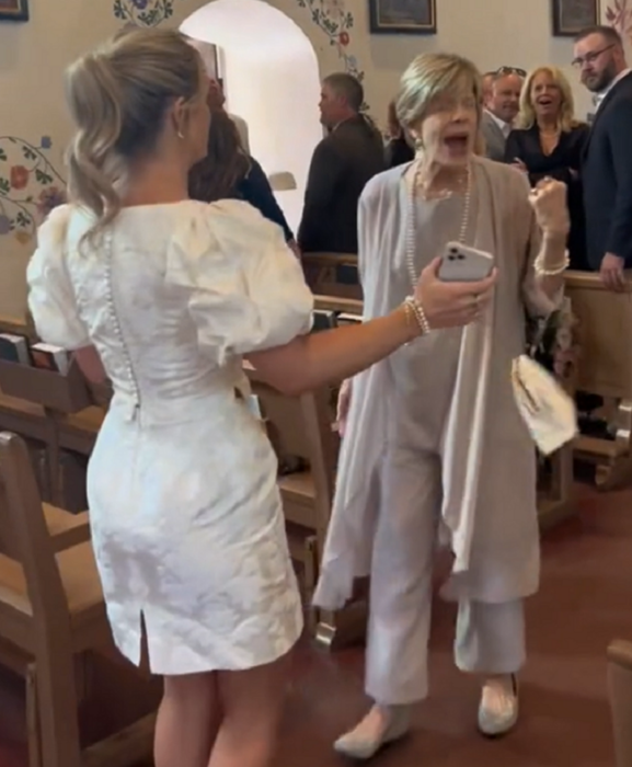 dentro de un templo una chica con vestido blanco espera a una mujer mayor que está gritando sorprendida ambas están paradas sobre el pasillo central