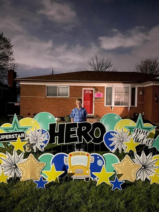 un chico caucásico posa en el jardín exterior de su casa junto a distintos carteles de colores que le colocaron sus vecinos en el pasto del frente de la vivienda 