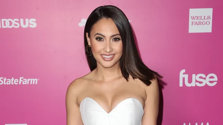 imagen de Francia Raisa en un evento público donde aparece vestida de blanco sobre una pared rosa con logotipos publicitarios