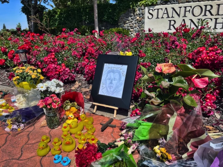 un altar lleno de flores con el dibujo del rostro de un hombre enmarcado en negro hay patitos de hule en el piso