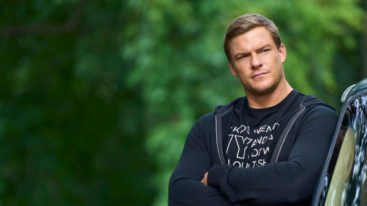 Alan Ritchson en una imagen donde luce cruzado de brazos recargado en un automóvil lleva ropa deportiva oscura 