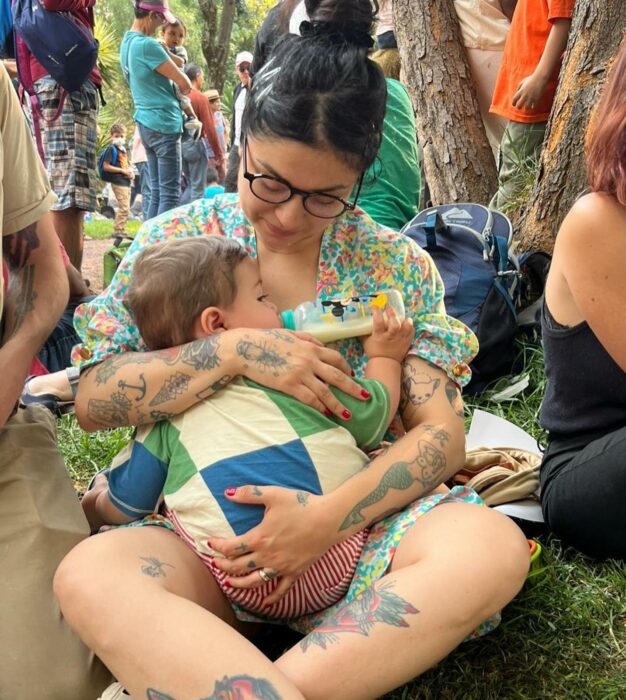 Mon Laferte sentada en el suelo cargando a su hijo Joelito mientras toma biberón. 