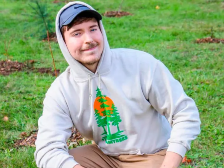 Mr Beats appears squatting, he is in the field as a green grass can be seen behind him, he is wearing a cap and has the hood of his sweatshirt on