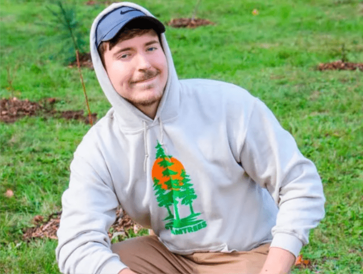 Mr Beats aparece en cuclillas está en el campo pues se ve un césped verde atrás de él lleva gorra y tiene puesta la capucha de la sudadera