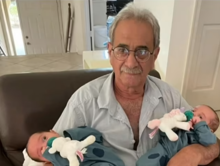 un hombre mayor carga en brazos a dos bebes que parecen ser gemelos