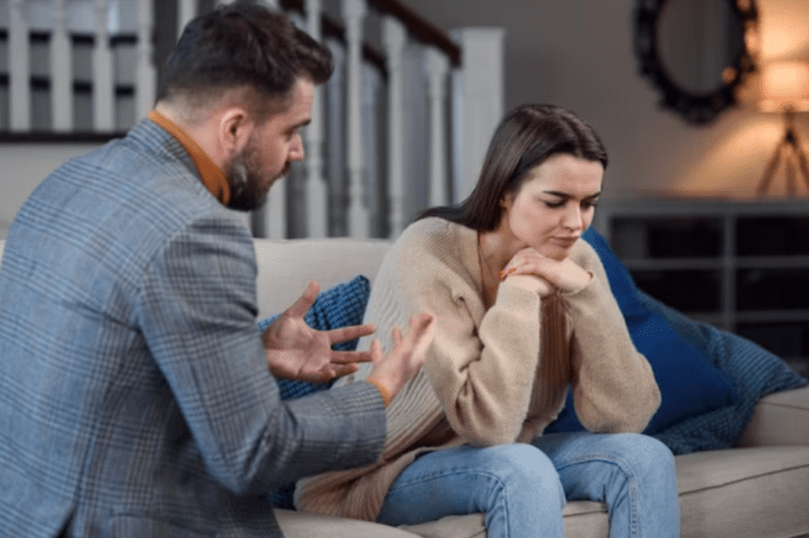 una pareja tiene una discusión en la sala de su casa ella tiene la vista en el piso y las manos sobre la barbilla él está hablando usando sus manos