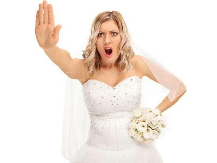 una novia con expresión de rechazo en el rostro levanta la mano como negándole el paso a alguien lleva un ramo en la otra mano