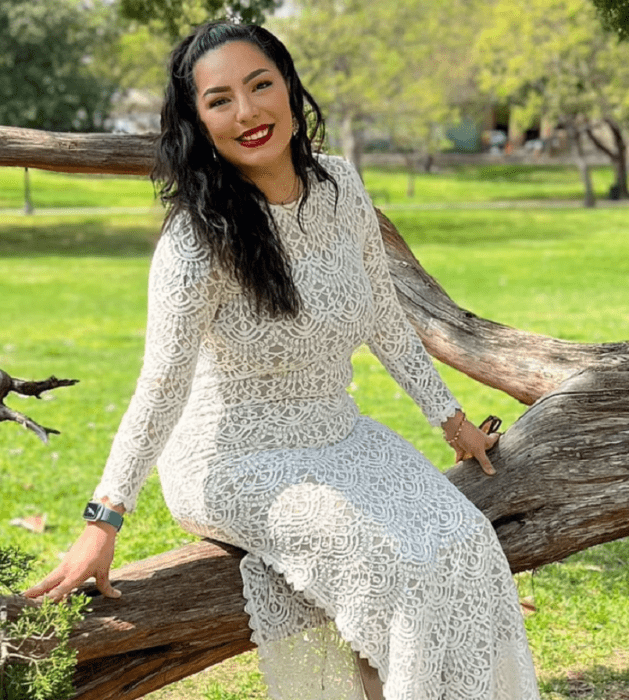 una mujer posa sentada en el tronco de un árbol que está paralelo al suelo lleva un vestido largo blanco