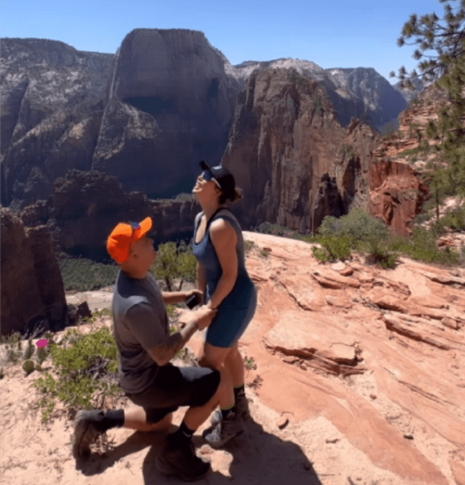 Emilio Sánchez hincado proponiéndole matrimonio a Chiquis Rivera en la cima de una montaña ambos llevan ropa deportiva