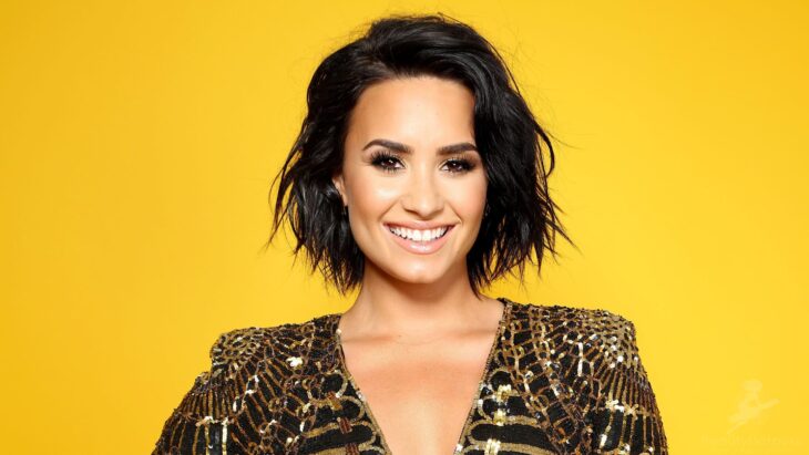 Demi Lovato posa sonriente con un fondo amarillo detrás lleva un vestido dorado con negro