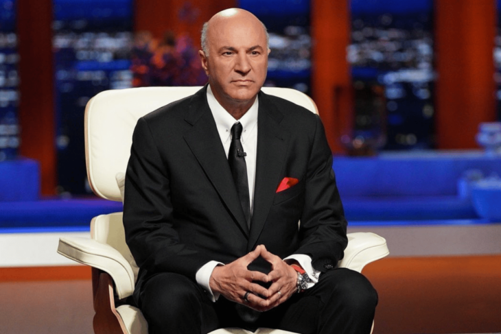 Kevin O'Leary se toca la punta de los dedos de las manos al frente mientras está sentado en uan enorme silla de color blanco lleva un traje con corbata negros
