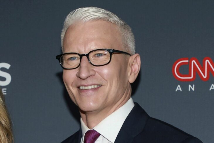 Anderson Cooper sonríe en algún evento lleva anteojos con armazón de color negro luce el cabello canoso y un traje negro con corbata guinda