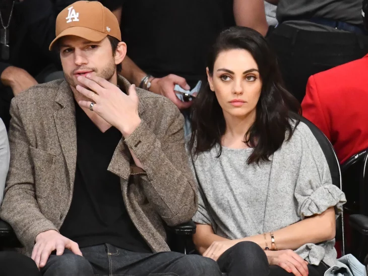 Ashton Kutcher y Mila Kunis están sentados en las gradas de un lugar deportivo él lleva gorra y se colocó una mano en la barbilla