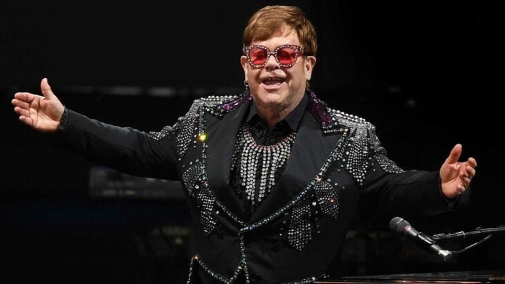 el cantante Elton John con un traje negro lleno de pedrería sobre el escenario luce unos anteojos con las micas de color rosa