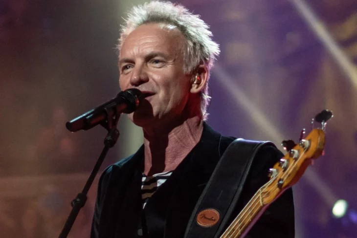 Sting está sobre el escenario trae una guitarra colgada al frente y está cantando en un micrófono con base el cabello luce rubio