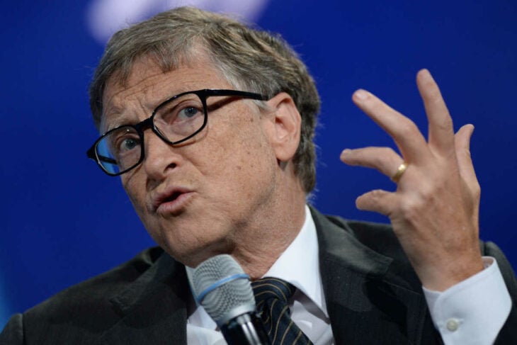 Bill Gates sostiene un micrófono mientras hace un ademán con la mano tiene un semblante preocupado y lleva anteojos