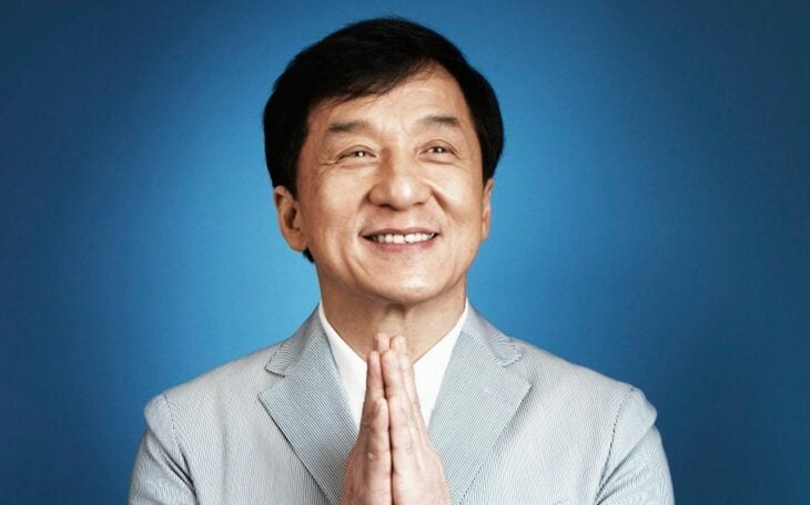 Jackie Chan sonríe divertido mientras junta sus manos al frente como si agradeciera lleva traje con camisa blanca 