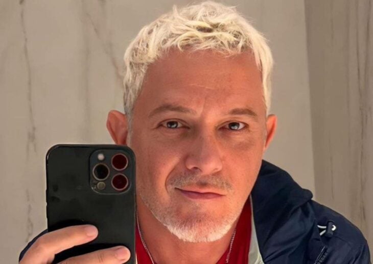 With a grimace, Alejandro Sanz poses in front of the mirror to take a selfie. He has blond hair and a slight white beard. 