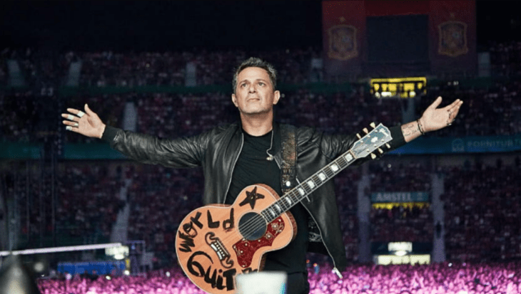 The singer Alejandro Sanz with a box guitar hanging on his chest opens his arms and looks at the sky while he is on stage