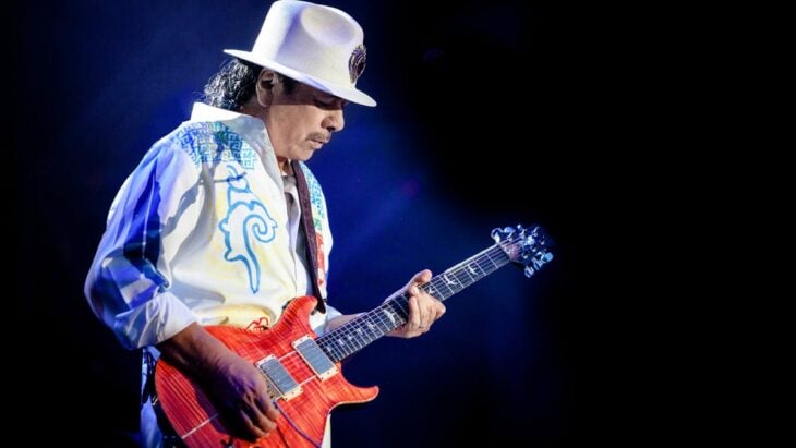 Carlos Santana vestido de blanco con una guitarra roja en sus manos está en el escenario tocando música