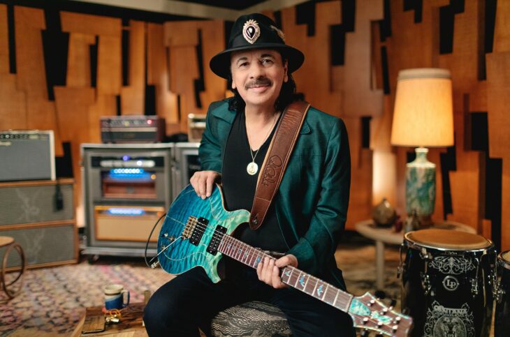 Carlos Santana posa sonriente con una guitarra verde esmeralda sentado en una habitación bajo la tenue luz de una lámpara 