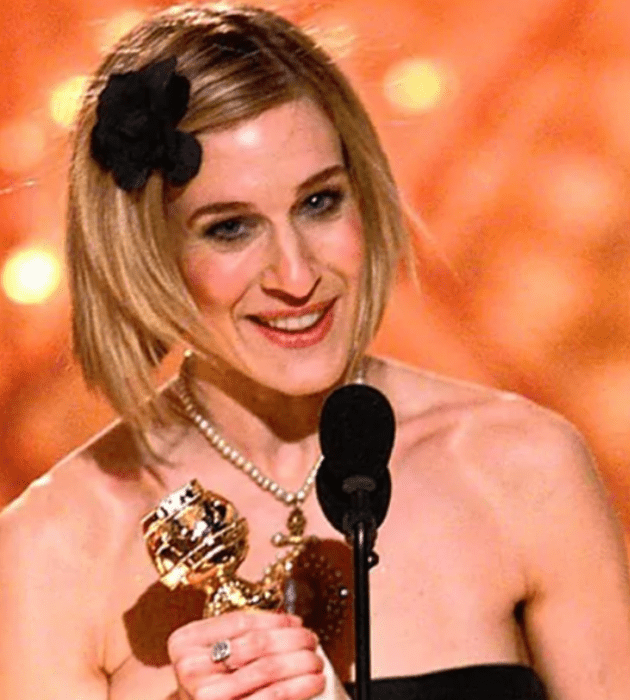 Sarah Jessica Parker al momento de recibir un premio que lleva en las manos está ante un micrófono y lleva un moño negro en el cabello