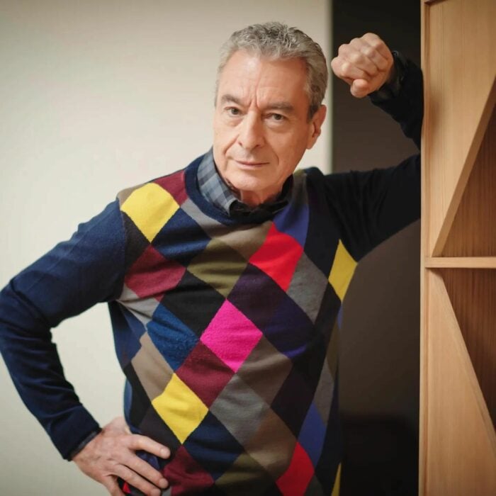 Fotografía del actor César Costa recargado en un librero vistiendo un suéter de rombos de colores 