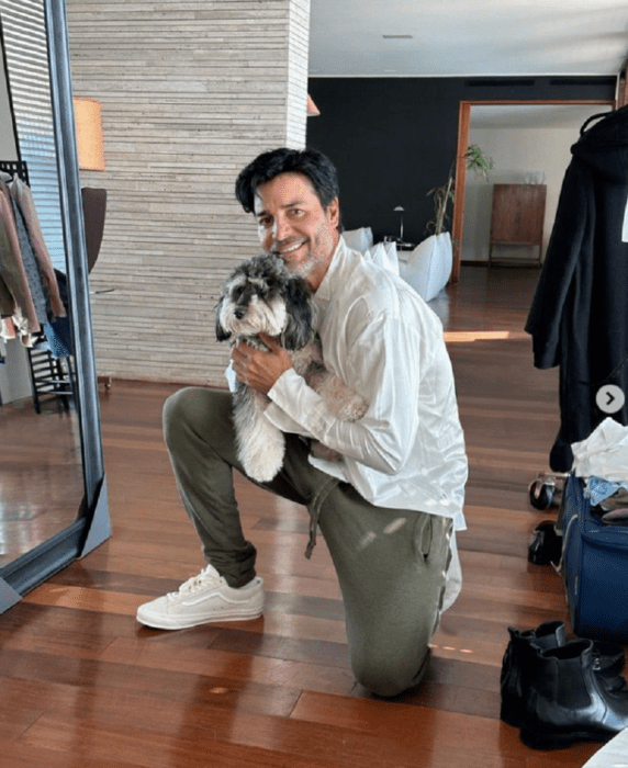 el cantante Chayanne está hincado sobre un piso de madera lleva en sus brazos un perrito de color blanco con negro