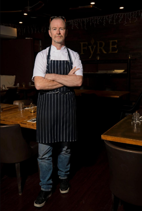 un chef con delantal negro está de brazos cruzados en su restaurante se le ve de cuerpo entero
