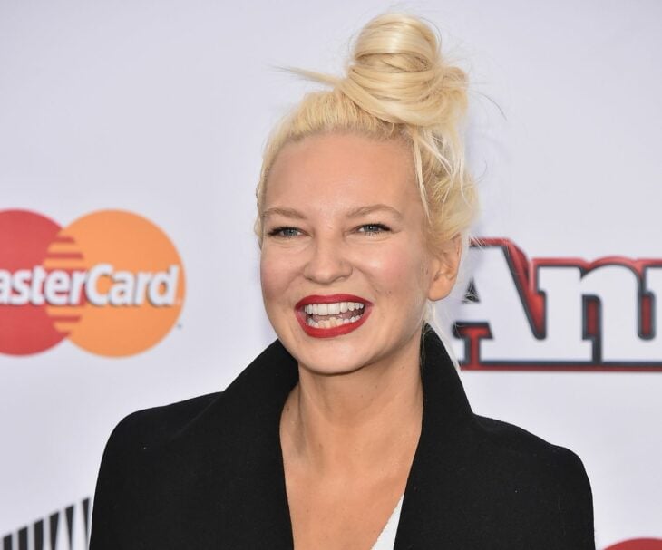 La cantante Sia posando en la alfombra roja de algún evento está sonriente y lleva el cabello rubio agarrado en un chongo despeinado 
