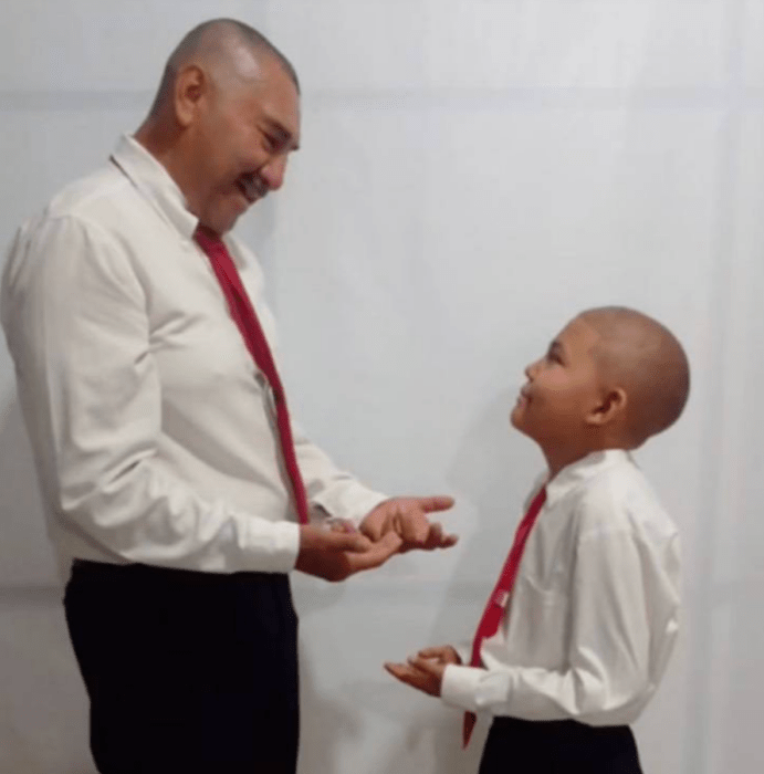 un hombre habla con un niño ambos están uniformados con camisa blanca y corbata roja llevan la cabeza rapada