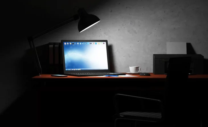 una computadora encendida en la oscuridad sobre un escritorio la alumbra una lámpara pequeña