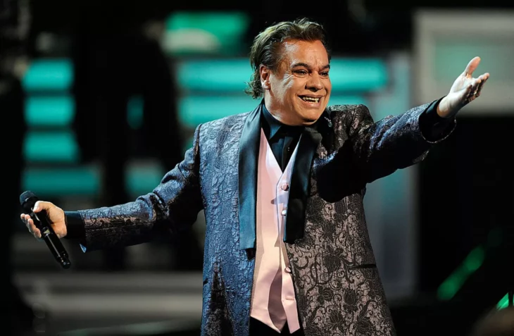 Juan Gabriel en concierto lleva un saco oscuro con chaleco blanco y sonríe mientras extiende los brazos