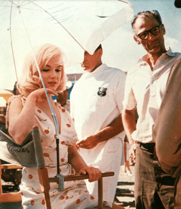 Marilyn Monroe sentada bajo una sombrilla en un descanso de la película Los inadaptados en la imagen aparece su esposo en ese momento Arthur Miller