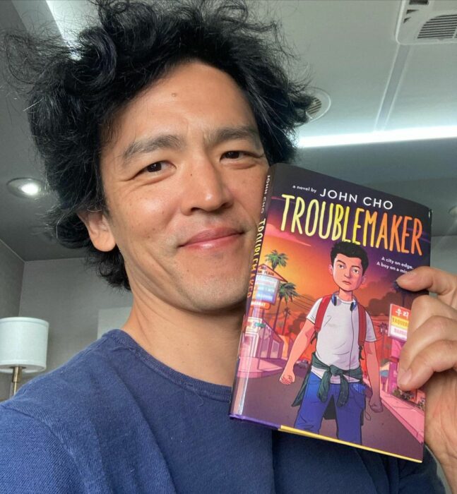 John Cho con playera azul mostrando un libro 