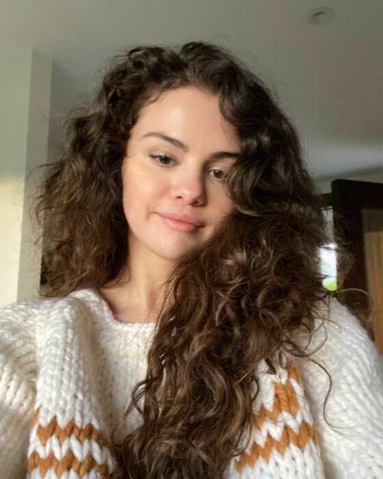 Selena Gomez con su melena suelta encrespada y sueter blanco 