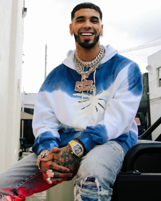 Anuel AA sentado con sudadera blanca con azul 