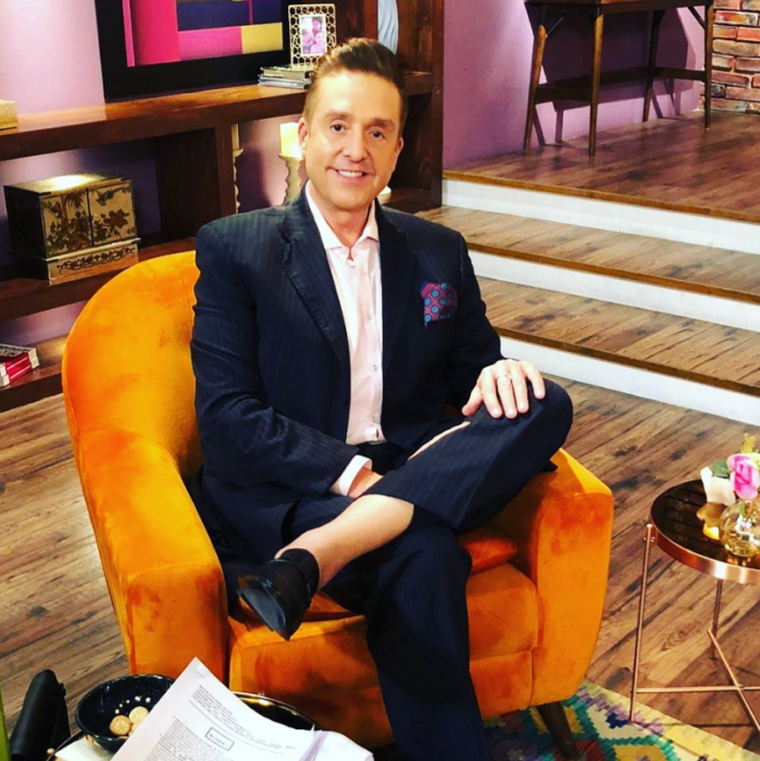 Daniel Bisogno posa sentado en un sillón naranja en el foro del programa Ventaneando lleva saco oscuro pero sin corbata