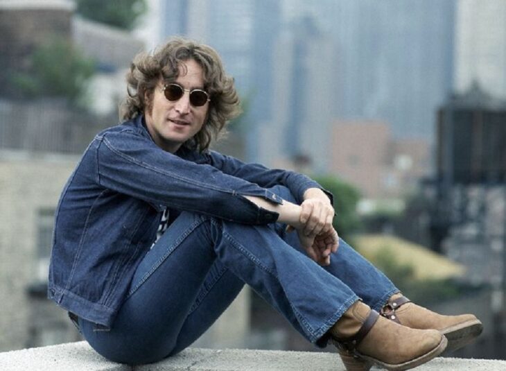 John Lennon posando sentado en la orilla de la barda de una terraza en Nueva York lleva ropa de mezclilla y lentes de sol