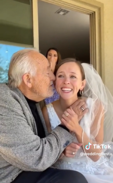un hombre mayor se acerca a besar la mejilla de su hija que está vestida de novia 