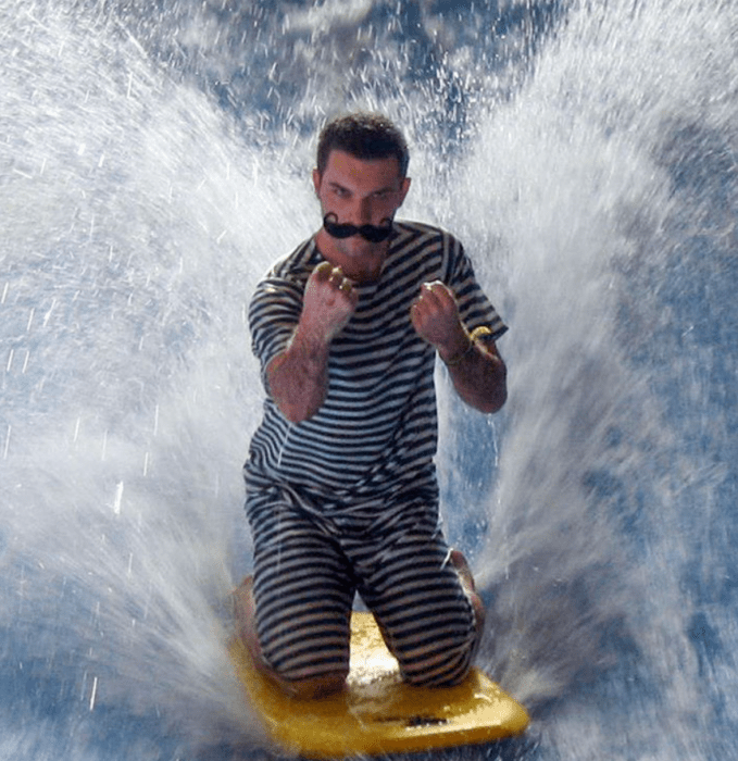 un hombre con traje antiguo y bigote extraño montado en una tabla de surf sobre una ola