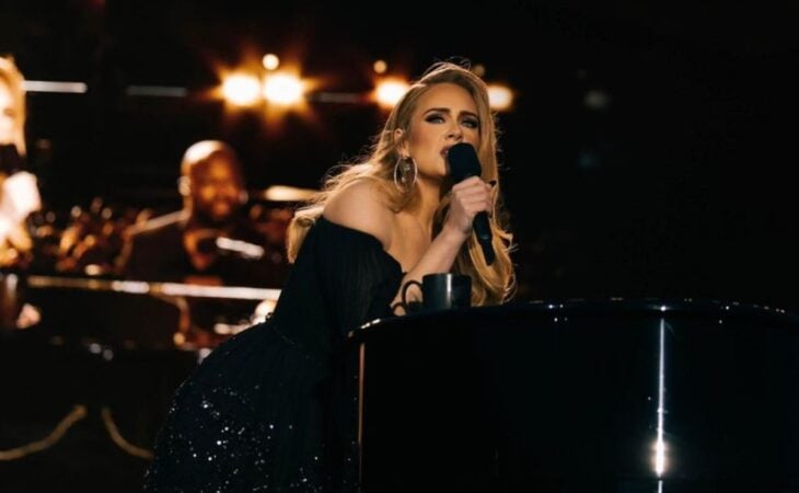 La cantante Adele durante uno de sus conciertos lleva un micrófono en la mano mientras se recarga en un piano negro cantando una canción