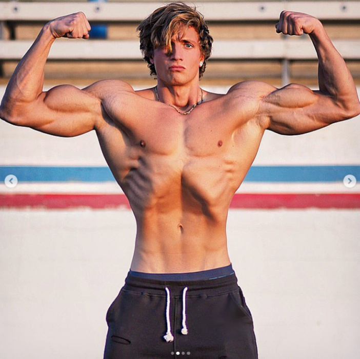 Joesthetics mostrando sus impresionantes músculos de fisiculturista con el torso desnudo y los brazos levantados