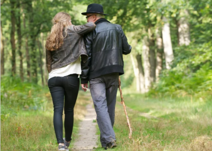 una hija camina con su papá por un camino llenó de vegetación el hombre lleva sombrero y bastón ella viste casual